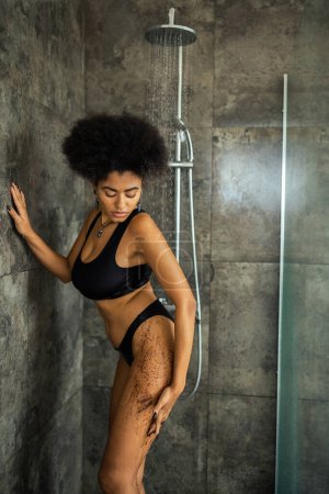 Curly african american woman applying coffee scrub under water in shower cabin