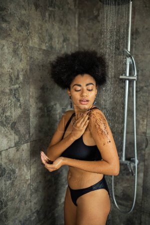 Pretty african american woman applying coffee scrub on shoulder in shower 