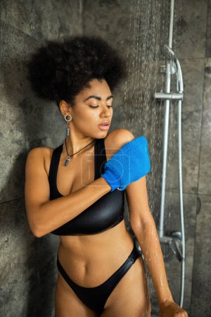 Téléchargez les photos : Jeune femme afro-américaine nettoyage du corps avec un gant de massage dans la douche - en image libre de droit