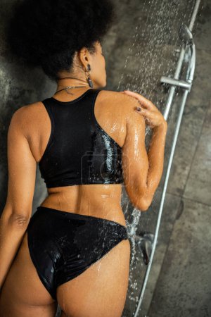Lockige Afroamerikanerin steht zu Hause unter Wasser in Duschkabine 