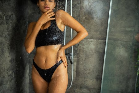 Ausgeschnittene Ansicht einer afrikanisch-amerikanischen Frau in Crop Top und Höschen unter der Dusche und mit Blick auf die Kamera zu Hause 