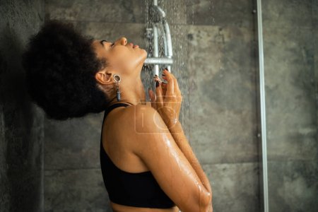 Téléchargez les photos : Vue latérale de la jeune femme afro-américaine en haut de culture prenant une douche à la maison - en image libre de droit