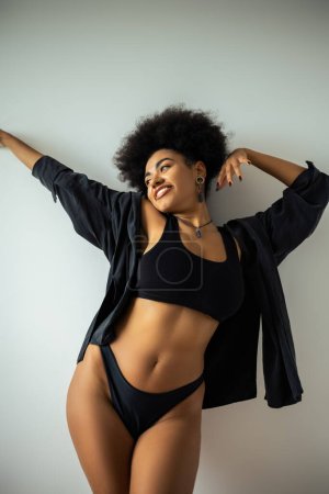 Femme afro-américaine joyeuse en chemise noire et sous-vêtements debout près du mur 