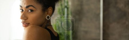 Curly african american woman looking at camera near glass of shower cabin at home, banner 