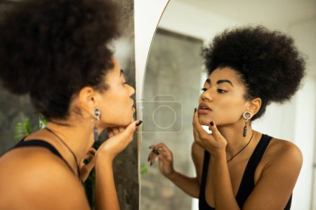 Widok z boku zamazanej afrykańskiej kobiety dotykającej twarzy podczas patrzenia w lustro w łazience