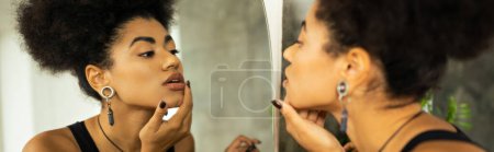 Widok z boku afrykańskiej kobiety dotykającej podbródka patrząc w lustro w domu, sztandar 