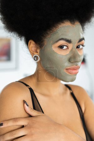 Téléchargez les photos : Portrait de femme afro-américaine avec masque en argile sur le visage regardant la caméra à la maison - en image libre de droit