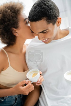 joven afroamericana mujer con taza de café diciendo secreto a sonriente novio en casa