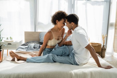 Foto de Seductora mujer afroamericana tocando barbilla de novio en jeans en la cama - Imagen libre de derechos