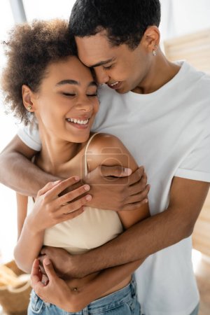 curly african american woman smiling with closed eyes near boyfriend hugging her at home
