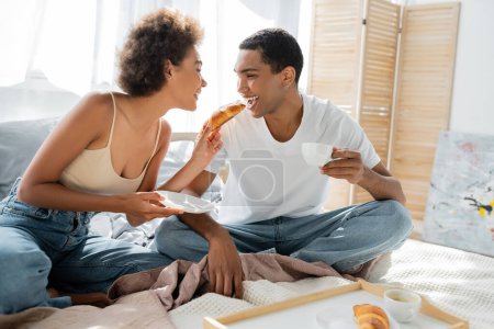 cheerful african american woman feeding boyfriend with croissant in bedroom