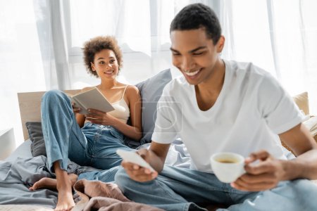 alegre afroamericana mujer sentada en la cama con libro y mirando novio con café y teléfono móvil en primer plano borrosa