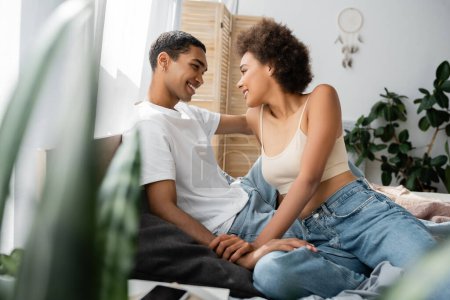 sexy african american couple sitting on bed and smiling at each other on blurred foreground