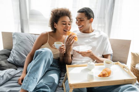 african american man in white t-shirt feeding girlfriend with croissant while having breakfast on bed
