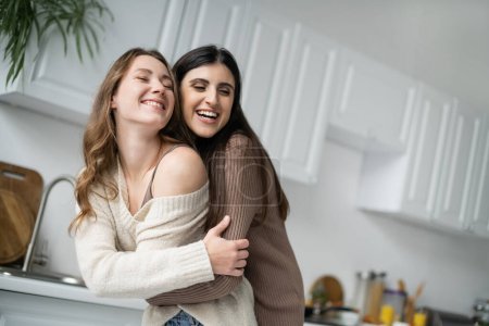 Cheerful lesbian woman hugging young girlfriend in kitchen 
