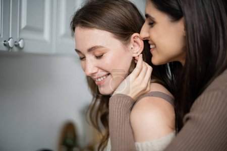 Positive woman touching ear of girlfriend in kitchen at home 