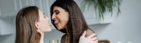 Young lesbian couple sticking out tongues in kitchen, banner 