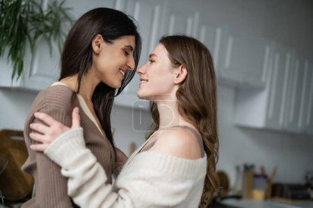 Photo for Side view of cheerful lesbian women hugging in blurred kitchen - Royalty Free Image