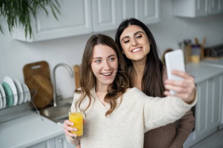 Téléchargez les photos : Couple lesbien souriant avec jus d'orange prenant selfie sur smartphone dans la cuisine - en image libre de droit