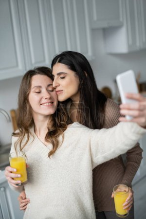 Photo for Lesbian woman holding orange juice and kissing girlfriend while taking selfie in kitchen - Royalty Free Image