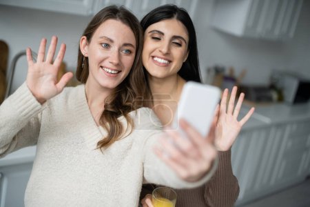 Téléchargez les photos : Sourire couple lesbien agitant les mains lors d'un appel vidéo sur smartphone dans la cuisine - en image libre de droit