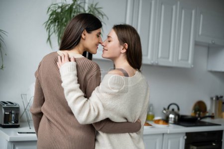 Téléchargez les photos : Vue latérale d'un couple lesbien souriant embrassant et se tenant nez à nez dans la cuisine - en image libre de droit