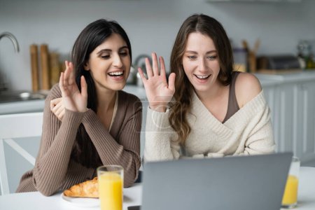 Positive lesbian couple having video call on laptop near breakfast in kitchen 