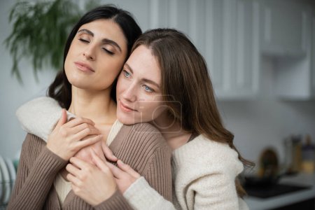 Young woman hugging lesbian partner with closed eyes in kitchen 