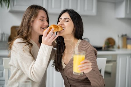 Young woman feeding girlfriend with croissant during breakfast in kitchen 