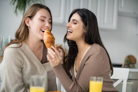 Positive lesbian woman holding croissant near girlfriend and glasses of orange juice in kitchen 