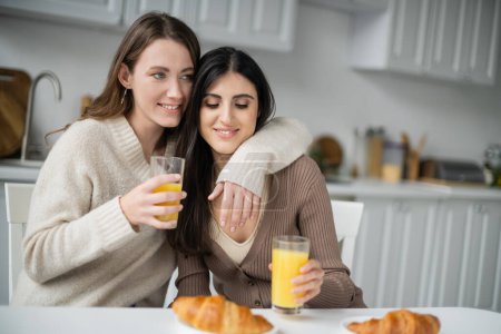 Positive woman hugging girlfriend near blurred croissant and orange juice in kitchen 