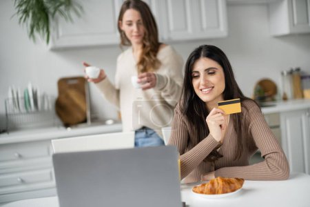 Téléchargez les photos : Femme souriante tenant carte de crédit près d'un ordinateur portable et petite amie floue pendant le petit déjeuner dans la cuisine - en image libre de droit