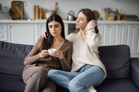 Lesbian woman hugging upset girlfriend with cup of coffee on couch at home 