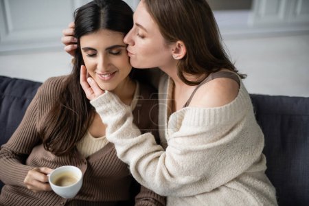Foto de Lesbiana mujer en caliente suéter besar novia con taza de café en sofá - Imagen libre de derechos