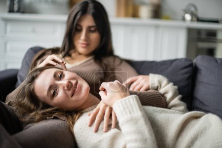 Téléchargez les photos : Jeune femme souriant à la caméra près floue lesbienne partenaire sur canapé - en image libre de droit