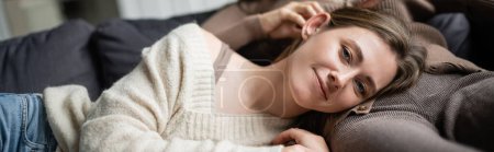 Foto de Mujer sonriente en suéter mirando a la cámara mientras está acostada cerca de su novia en el sofá, pancarta - Imagen libre de derechos