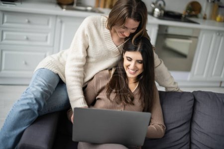 Photo for Cheerful lesbian couple in warm clothes using laptop on couch - Royalty Free Image