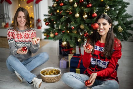 Cheerful woman in sweater throwing christmas ball near girlfriend and spruce tree in living room 