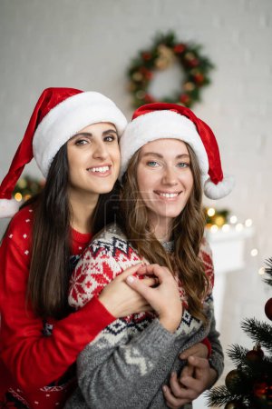 Foto de Feliz pareja lesbiana en sombreros de santa y suéteres tomados de la mano en casa - Imagen libre de derechos