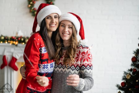 Happy same sex couple in santa hats holding sparklers and looking at camera at home 