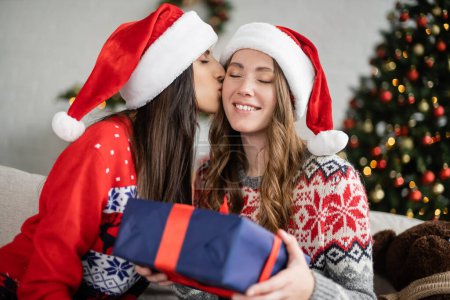 Lesbische Frau im Pullover küsst Freundin mit Weihnachtsmann-Hut und hält Geschenk zu Hause 