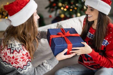 Verschwommene Frau mit Weihnachtsmütze macht Freundin im Wohnzimmer Weihnachtsgeschenk 