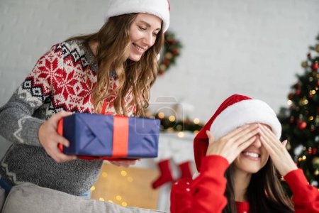 Smiling woman in santa hat holding blurred gift while girlfriend covering eyes at home 