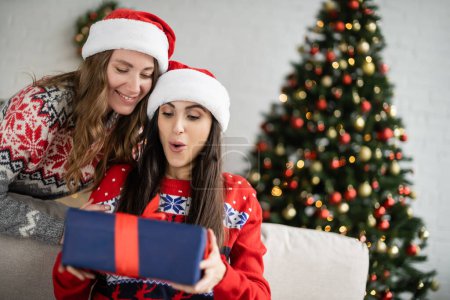 Lächelnde Frau mit Weihnachtsmütze schaut schockierte Freundin bei Neujahrsfeier mit Geschenk an 