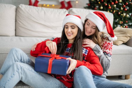 Lächelnde Frau mit Weihnachtsmütze umarmt Freundin mit Weihnachtsgeschenk im Wohnzimmer 