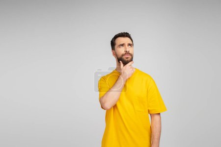Foto de Hombre pensativo en camiseta amarilla tocando la barba y mirando hacia otro lado aislado en gris - Imagen libre de derechos