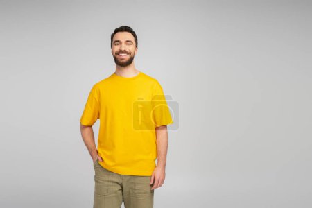 happy bearded man in yellow t-shirt standing with hand in pocket of beige trousers isolated on grey