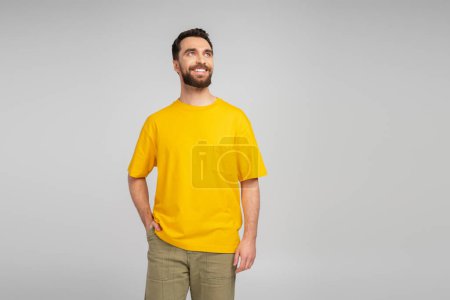 smiling bearded man standing with hand in pocket and looking away isolated on grey