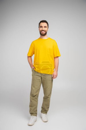 Foto de Longitud completa de hombre barbudo feliz en pantalones beige y camiseta amarilla de pie con la mano en el bolsillo sobre fondo gris - Imagen libre de derechos