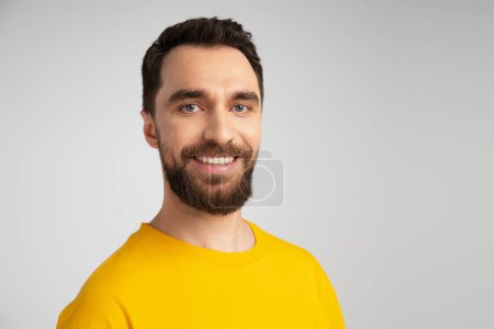 portrait of joyful bearded man in yellow t-shirt looking at camera isolated on grey